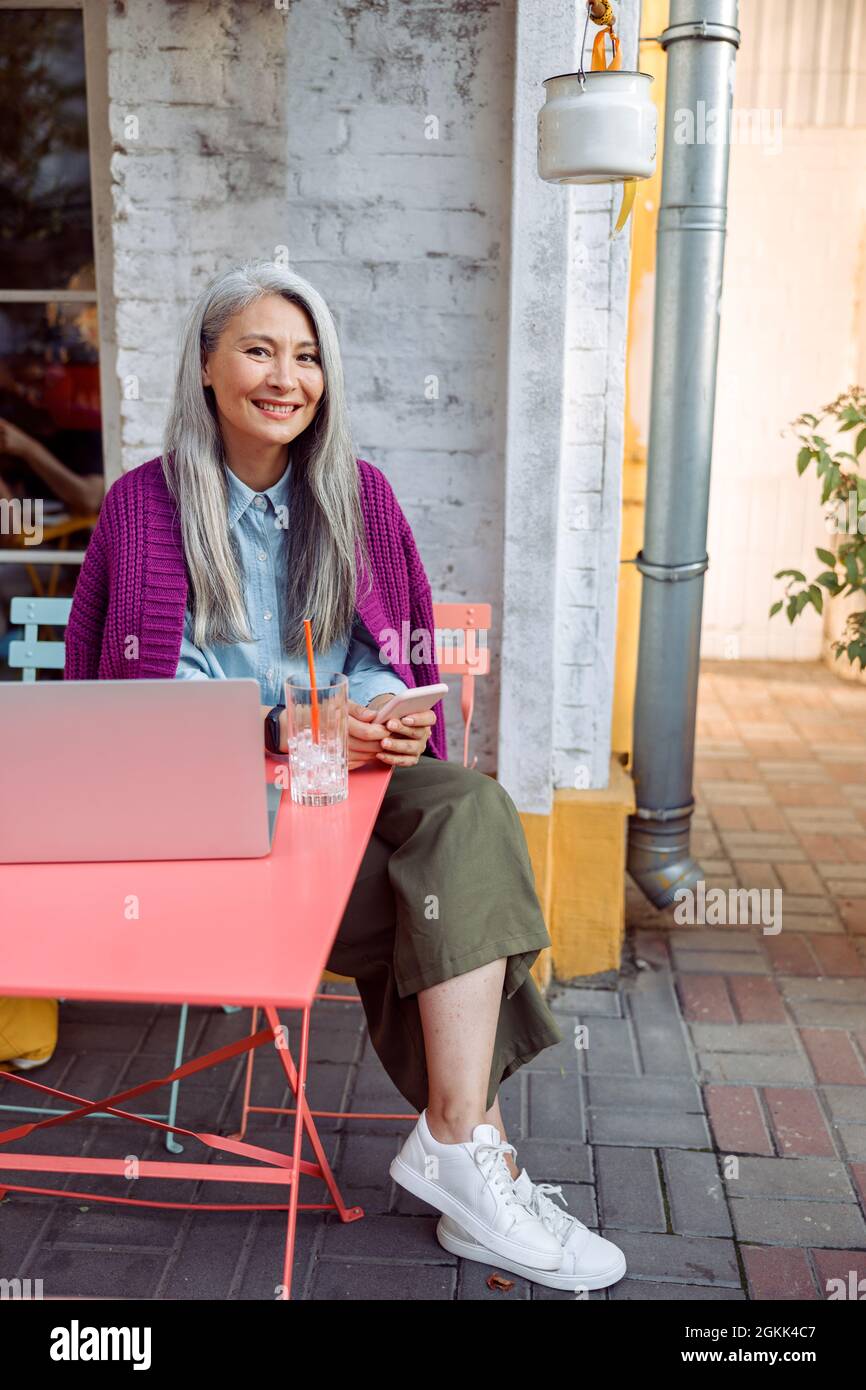 Lächelnde reife asiatische Dame in warmer purpurner Jacke hält Telefon am Tisch mit Laptop sitzen Stockfoto