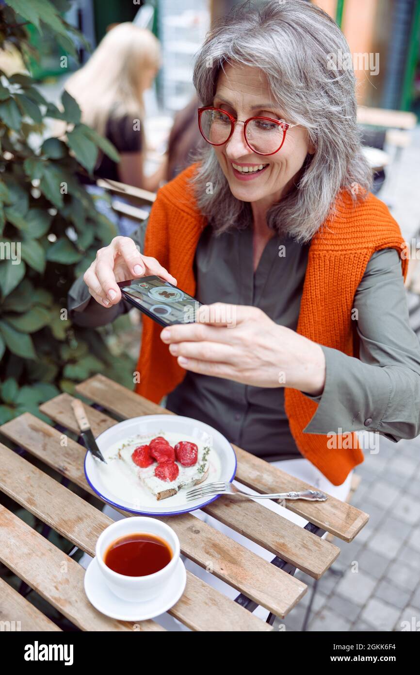 Positive ältere Frau fotografiert köstliche Erdbeerdessert auf der Terrasse des Cafés im Freien Stockfoto