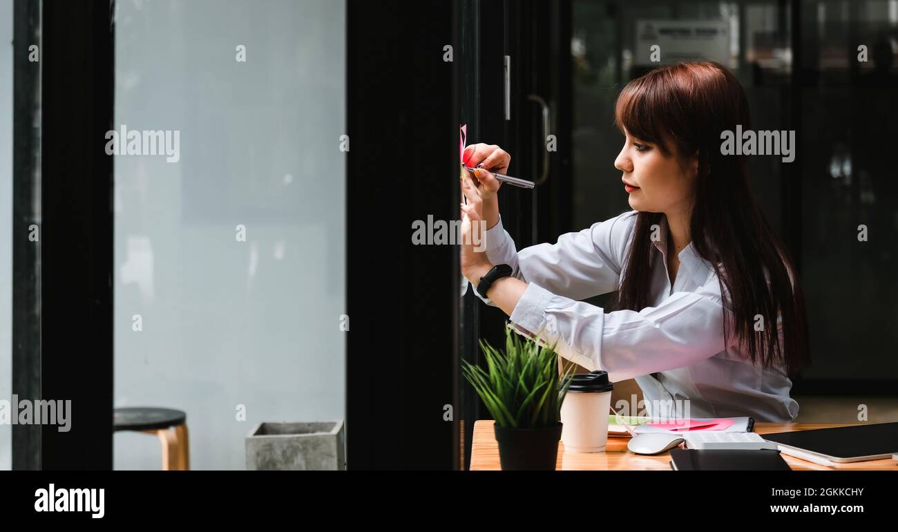Aufnahme einer asiatischen Frau, die während der Arbeit mit Finanzdienstleisterin in ihrem Büro Haftnotiz auf Glasfenster schrieb Stockfoto
