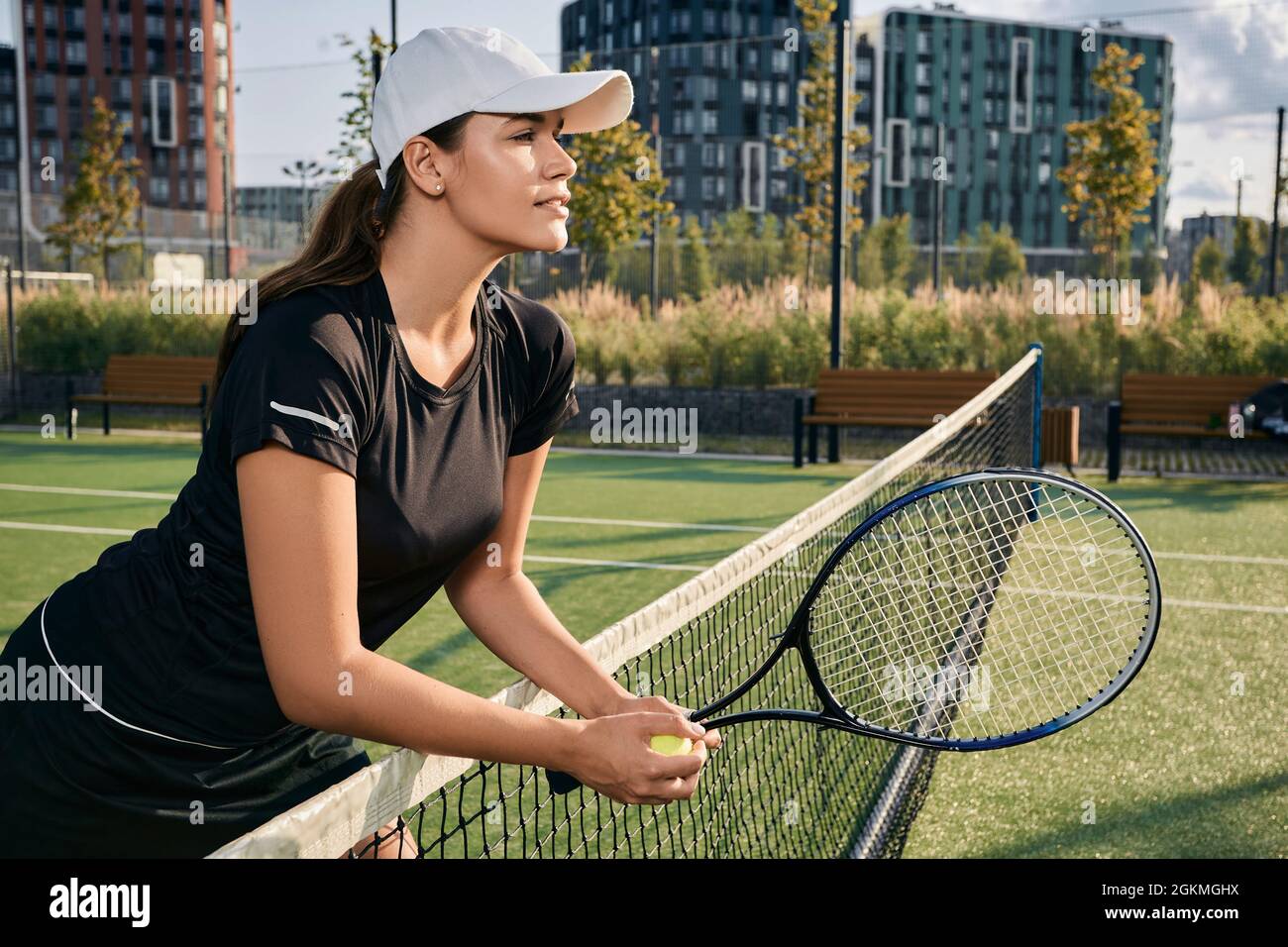 Tennisspielerin in Sonnenkappe lehnt sich auf das Netz, während sie auf dem Platz steht und Tennisschläger und -Ball hält, schaut in die Ferne auf ihre Gegnerin im Tennisspiel Stockfoto