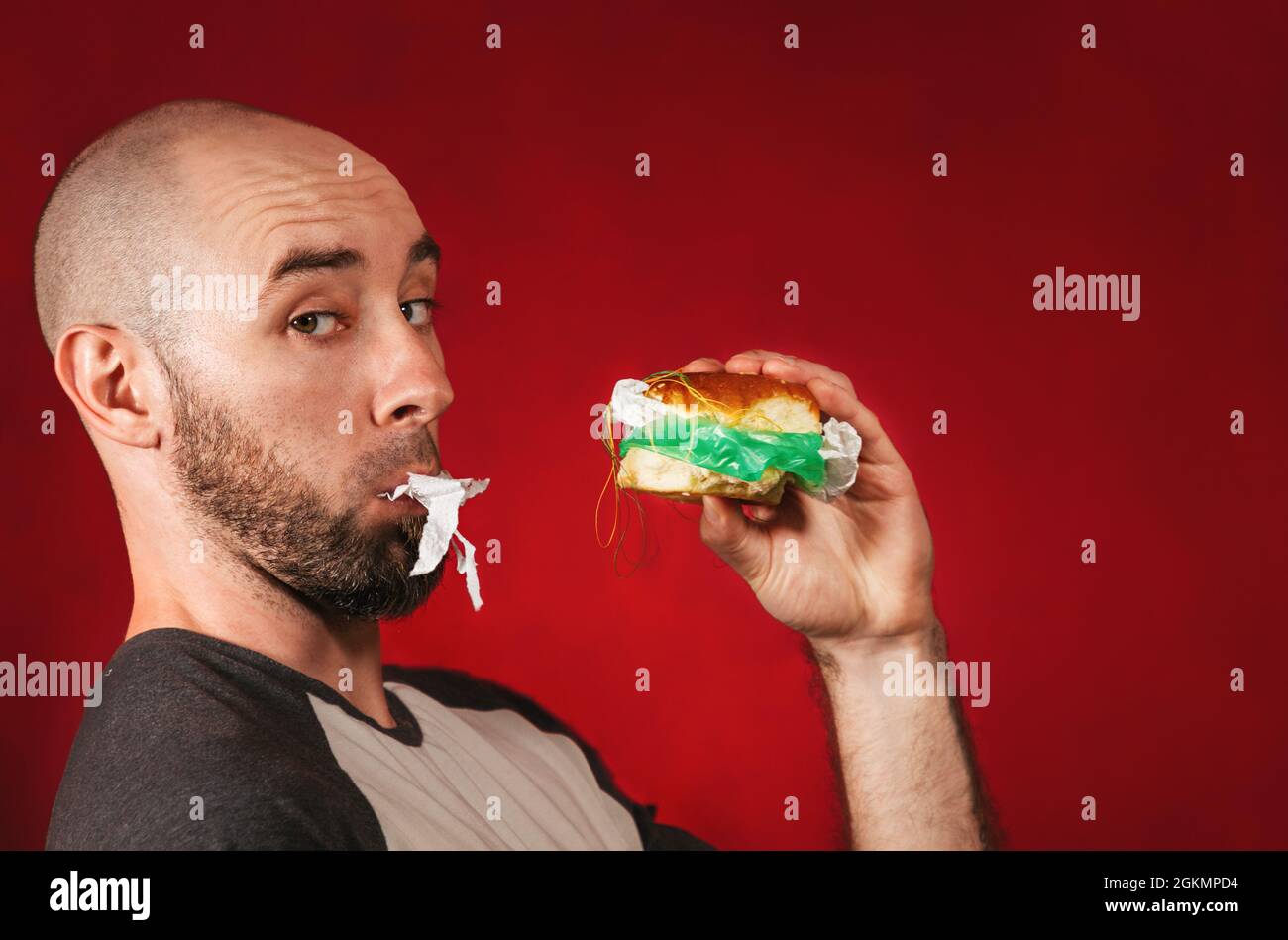 Umweltverschmutzung und Lebensmittelindustrie. Porträt eines kahlen Mannes mit Bart, der in einen mit Müll und Tüten gefüllten Hamburger beißt. Roter Hintergrund Stockfoto