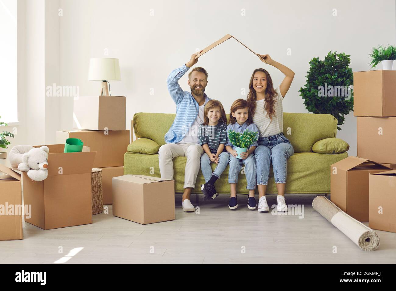 Glückliche Eltern, die das Dach über ihren Kindern halten, während sie im neuen Kinderhaus auf dem Sofa sitzen Stockfoto