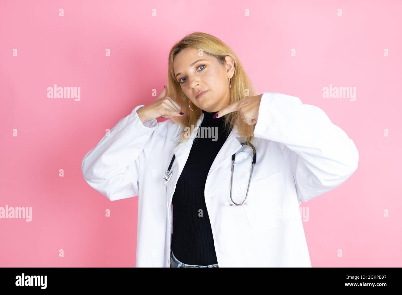 Junge blonde Ärztin, die Stethoskop trägt und auf einem isolierten rosa Hintergrund steht und die „Call me“-Geste mit ihren Händen macht. Stockfoto