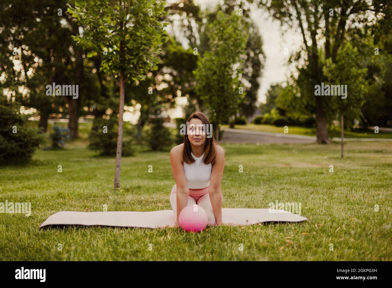 Charmante fit junge Frau sitzt auf den Knien auf Sportmatte mit Gummi kleinen Pilates Ball im Park Stockfoto