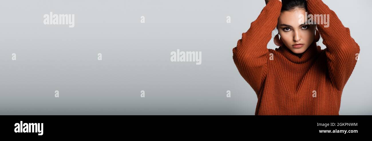 Model Frau in Ohrringen und Strickpullover Blick auf Kamera isoliert auf grauem Banner Stockfoto