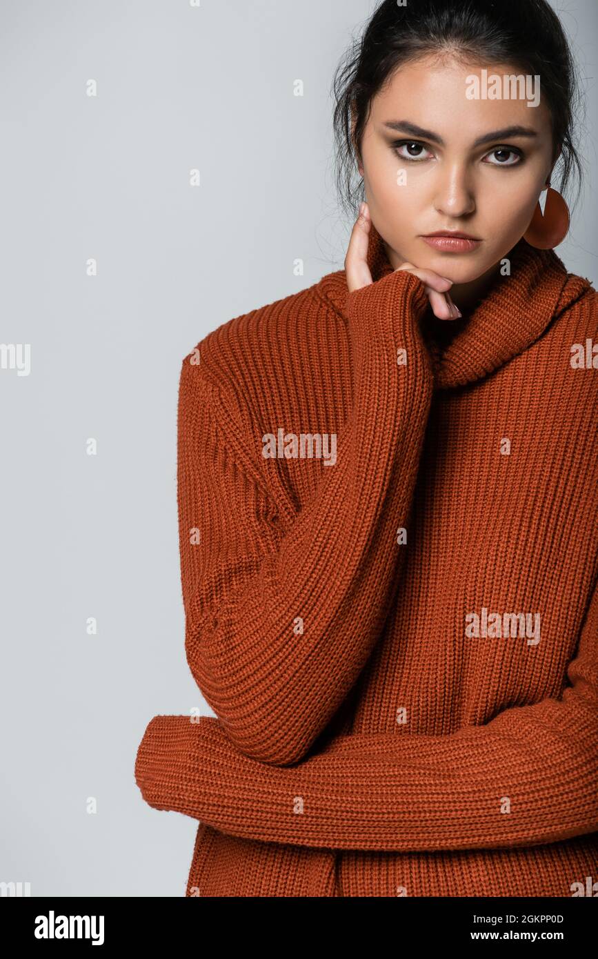 Junges Modell in Ohrringen und Strickpullover mit Blick auf die Kamera isoliert auf grau Stockfoto
