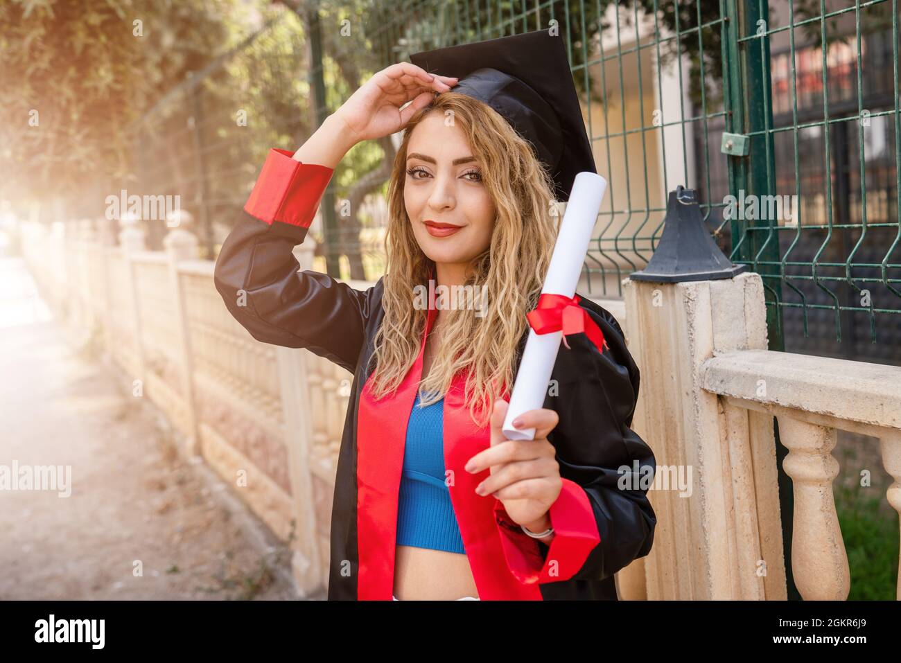 Eine hübsche Schülerin, die ihren Abschluss feiert und vor dem Schulzaun steht und ihr Diplom mit Erfolg hält. Hochwertige Fotos Stockfoto