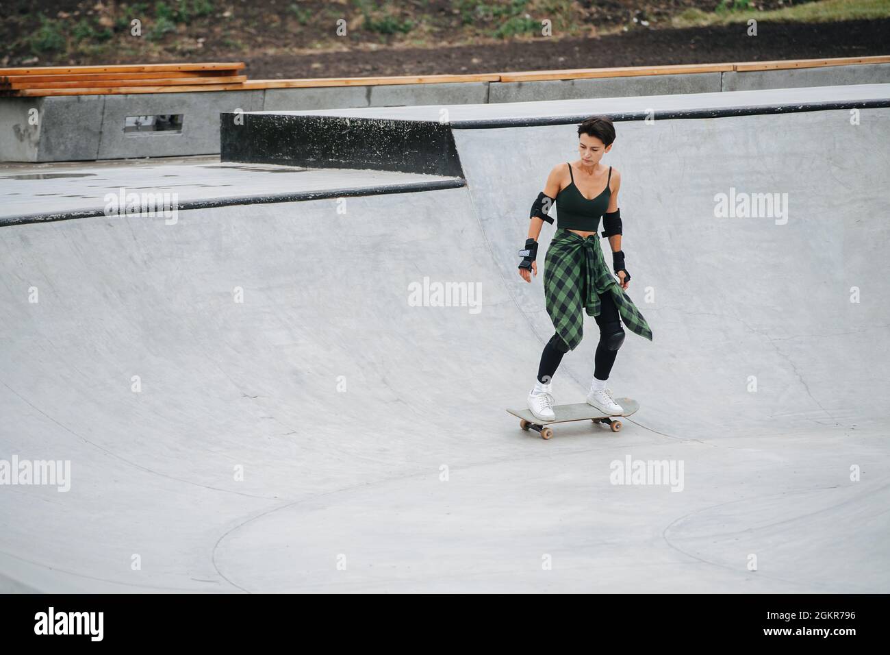 Longshot einer Skaterin, die auf ihrem Brett auf einem Viertelrohrkreis reitet. Sie ist in einer dunklen Kleidung mit weißen Sneakers getrauert. Stockfoto Longshot einer Skaterin, die auf ihrem Brett auf einem Viertelrohrkreis reitet. Sie ist in einer dunklen Kleidung mit weißen Sneakers getrauert. Stockfoto