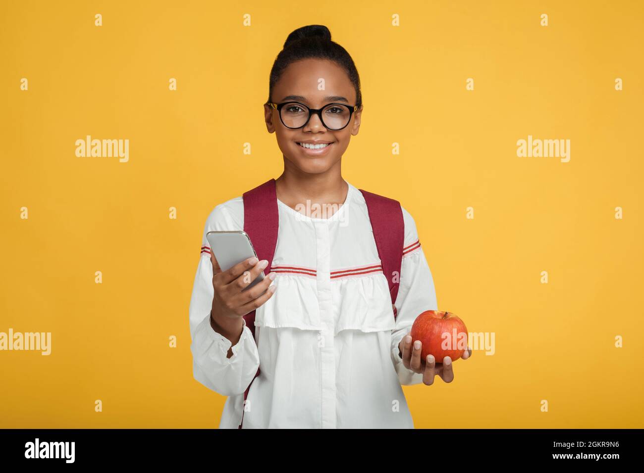 Happy smart junge afro amerikanische Mädchen Schüler in einer Brille mit Rucksack halten Smartphone und roten Apfel Stockfoto