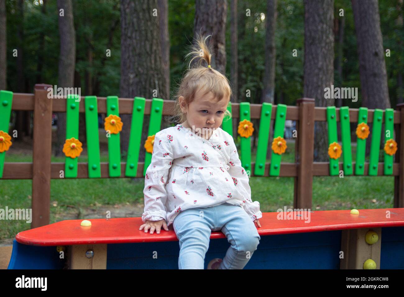 Nette liebenswert kaukasischen kleinen Mädchen spielt auf dem Spielplatz im Freien. Mädchen in einem weißen Kleid und Haar in einem Pferdeschwanz und einem traurigen Gesicht geflochten Stockfoto