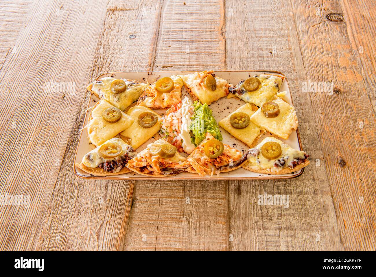 Version von Nachos mit Guacamole mit viel geschmolzenem Käse in jeder Portion einer Weizenmehl-Tortilla mit Jalapenos und auf einem hellen Holztisch Stockfoto