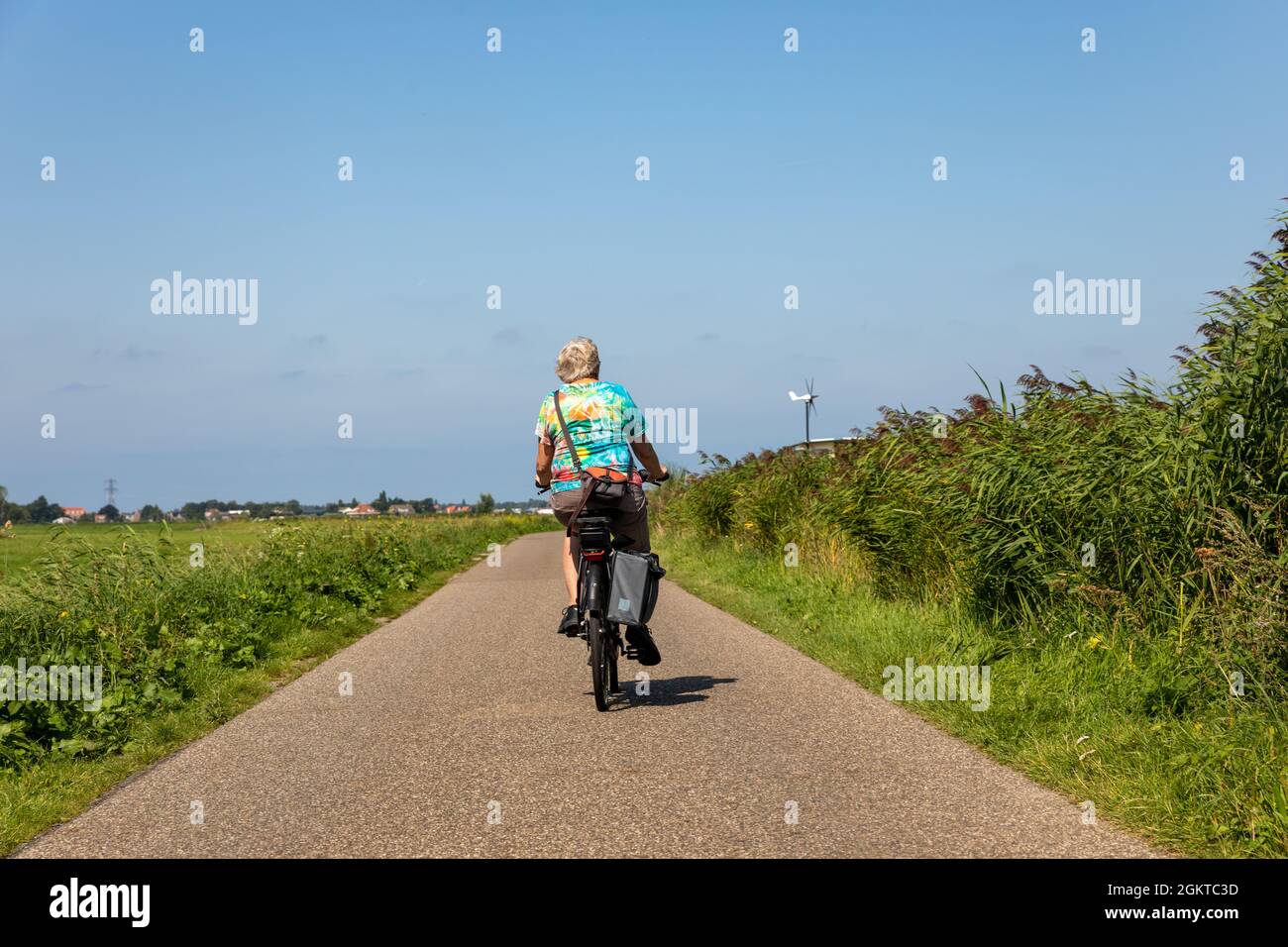 Ältere Dame, die auf einer asphaltierten Straße (Deich) mit einem Elektrofahrrad im südholländischen Dorf Lisse in den Niederlanden unterwegs ist. Von hinten aufgenommen. Mit einem Stockfoto