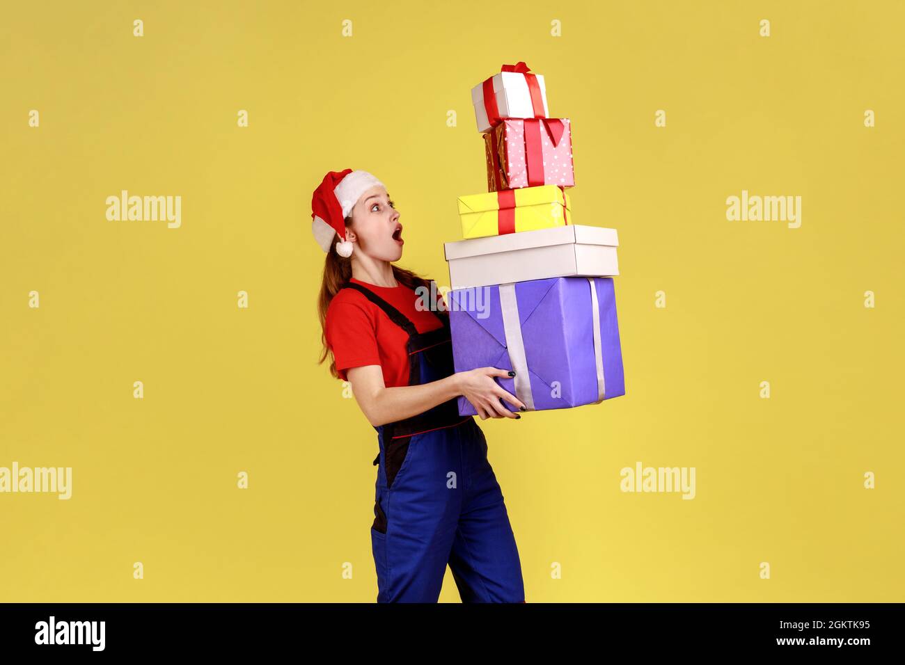 Aufgeregt Kurierin weiblich hält Stapel von Geschenken, Lieferung von Geschenken für Kunden für Winterferien, trägt blauen Overalls und weihnachtsmann Hut. Innen Stockfoto