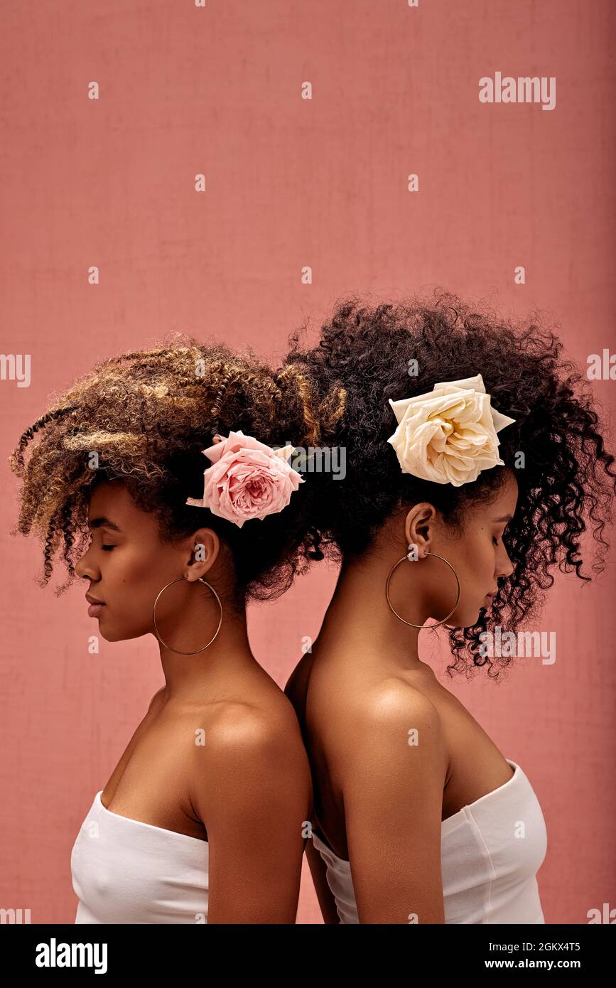 Zwei Frauen mit Blumen in den Haaren, die an einer rosa Wand mit geschlossenen Augen Rücken an Rücken stehen Stockfoto