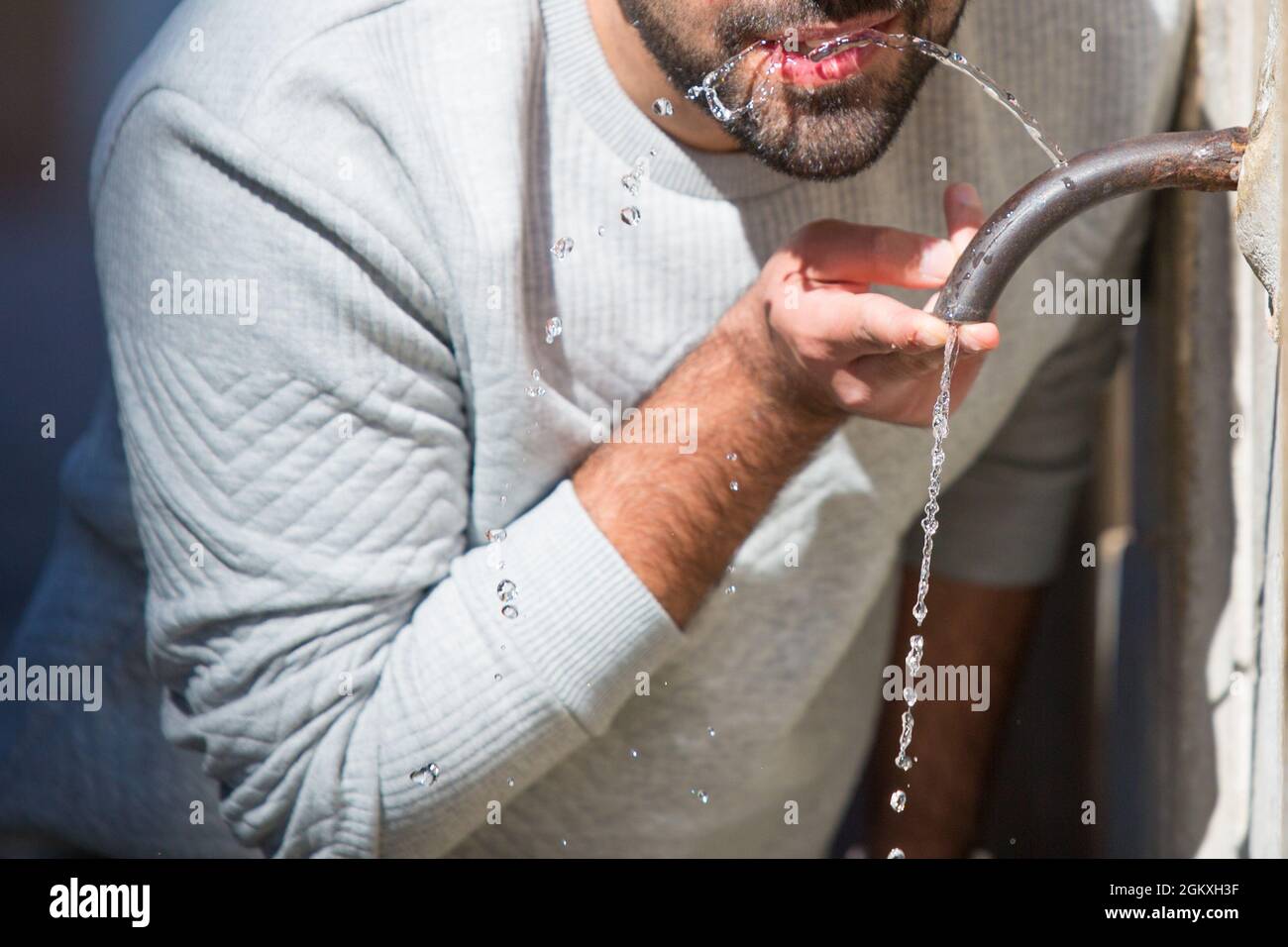 Der kaukasische Mann trinkt Wasser aus einem historischen traditionellen Trinkbrunnen im Freien in Rom. Heißer Sommertag. Lässiger weißer Pullover, blaue Jeans Stockfoto