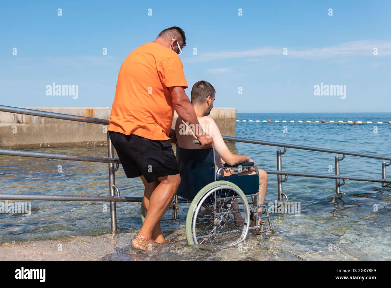 Behinderter Mann auf einem Rollstuhl, der zum Schwimmen mit einer Rampe ins Meer transportiert wurde. Stockfoto