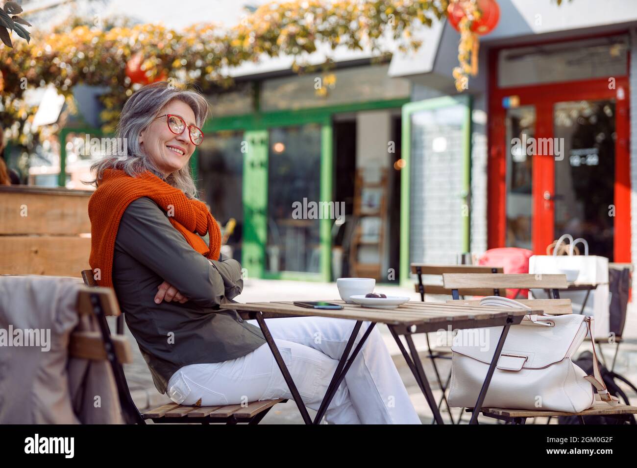 Eine nachdenkliche ältere Dame mit gekreuzten Armen sitzt am Tisch auf der Terrasse des Cafés Stockfoto