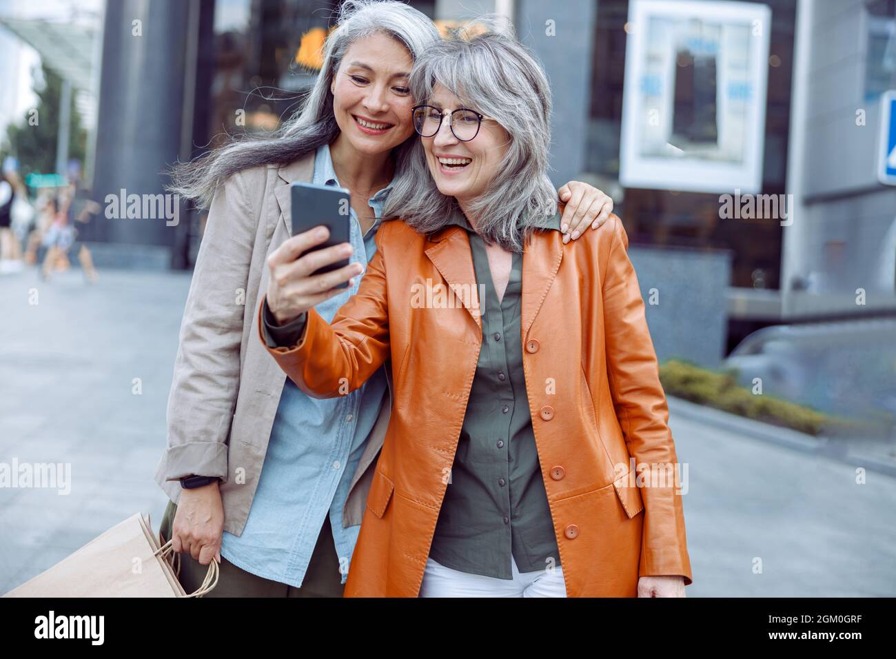 Lächelnde silberhaarige Dame und Begleiter mit Einkaufstaschen machen Selfie auf der modernen Stadtstraße Stockfoto