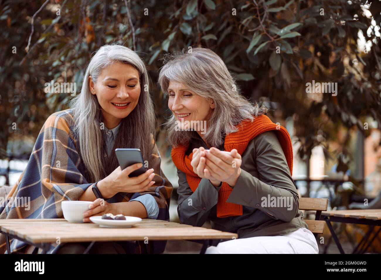 Lächelnde asiatische Dame zeigt einem Freund, der im Straßencafé sitzt, Fotos am Telefon Stockfoto