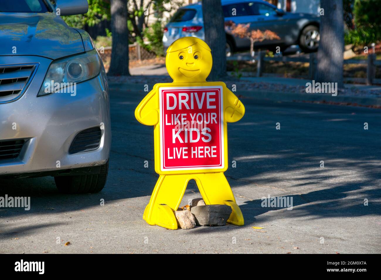 Fahren Sie wie Ihre Kinder Leben Sie hier Straßenschild in Wohnviertel in der Nähe geparkten Auto. Kindliche gelbe Figur mit rotem Sicherheitsschild auf der Straße. Stockfoto