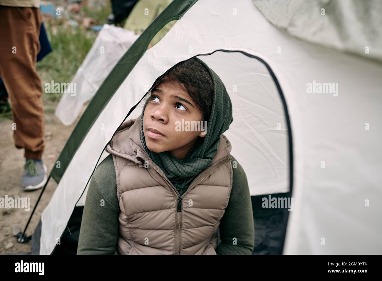 Ernsthafte obdachlose Flüchtling Middle-Eastern Mädchen in Kopftuch und Weste sitzen im Zelt und schauen hoffentlich auf Stockfoto