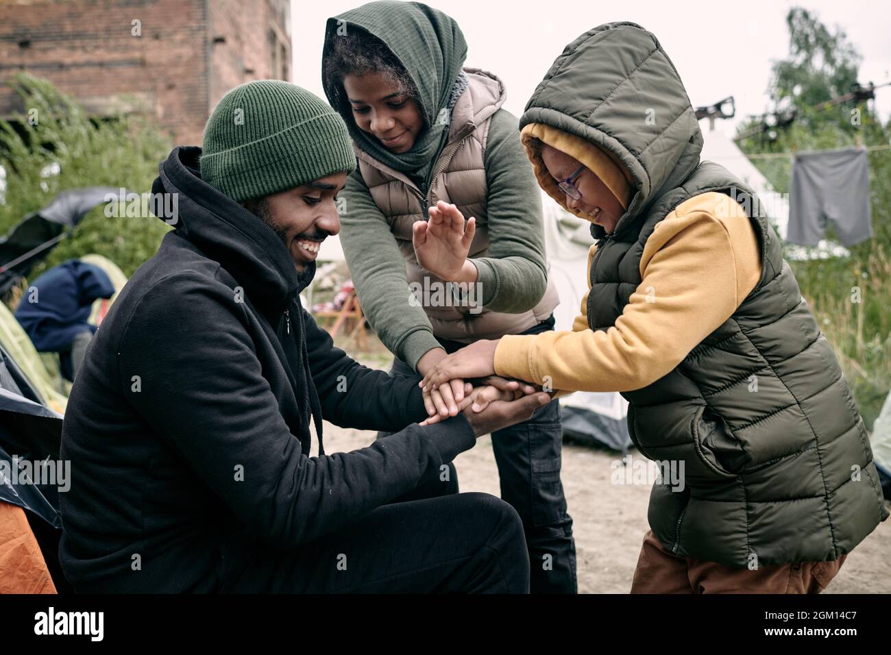 Positiver junger schwarzer Mann im Hut, der mit Kindern gegen verlassene Gebäude spielt und im Flüchtlingslager Kleidung an einem Seil hängt Stockfoto