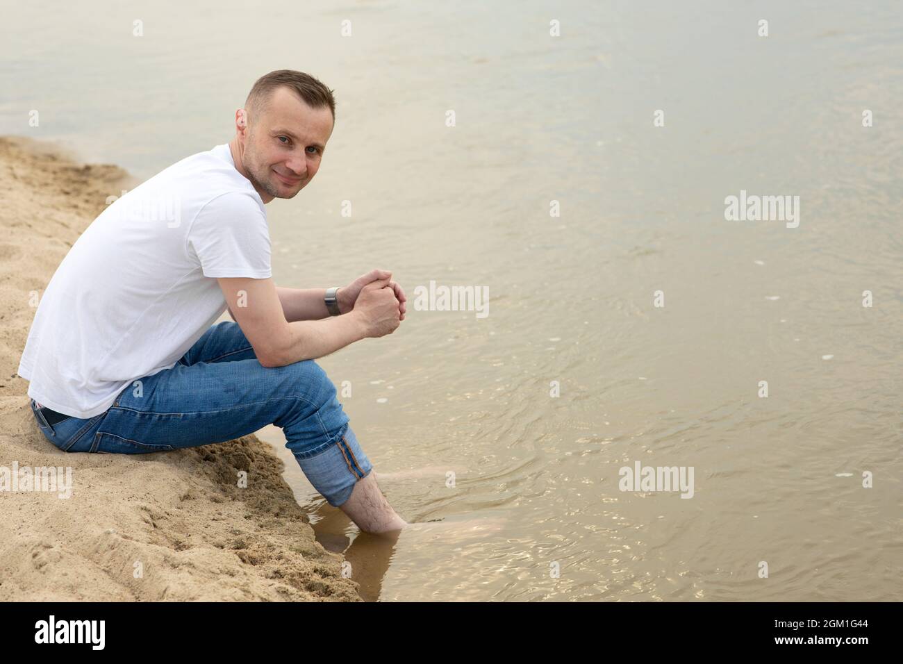 Das Bild einsam positiver und lächelnder Mann, der an einem Strand am Fluss sitzt und seine Beine im Wasser hält Stockfoto