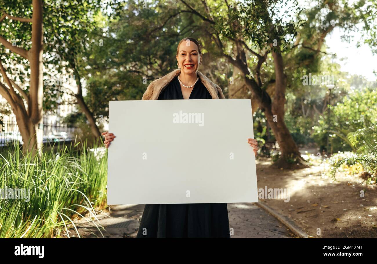 Fröhliche, schicke Frau, die die Kamera anlächelt, während sie ein weißes Plakat hochhält. Glückliche Frau mittleren Alters, die ein leeres Banner zeigt. Weibliche Aktivistin Standin Stockfoto