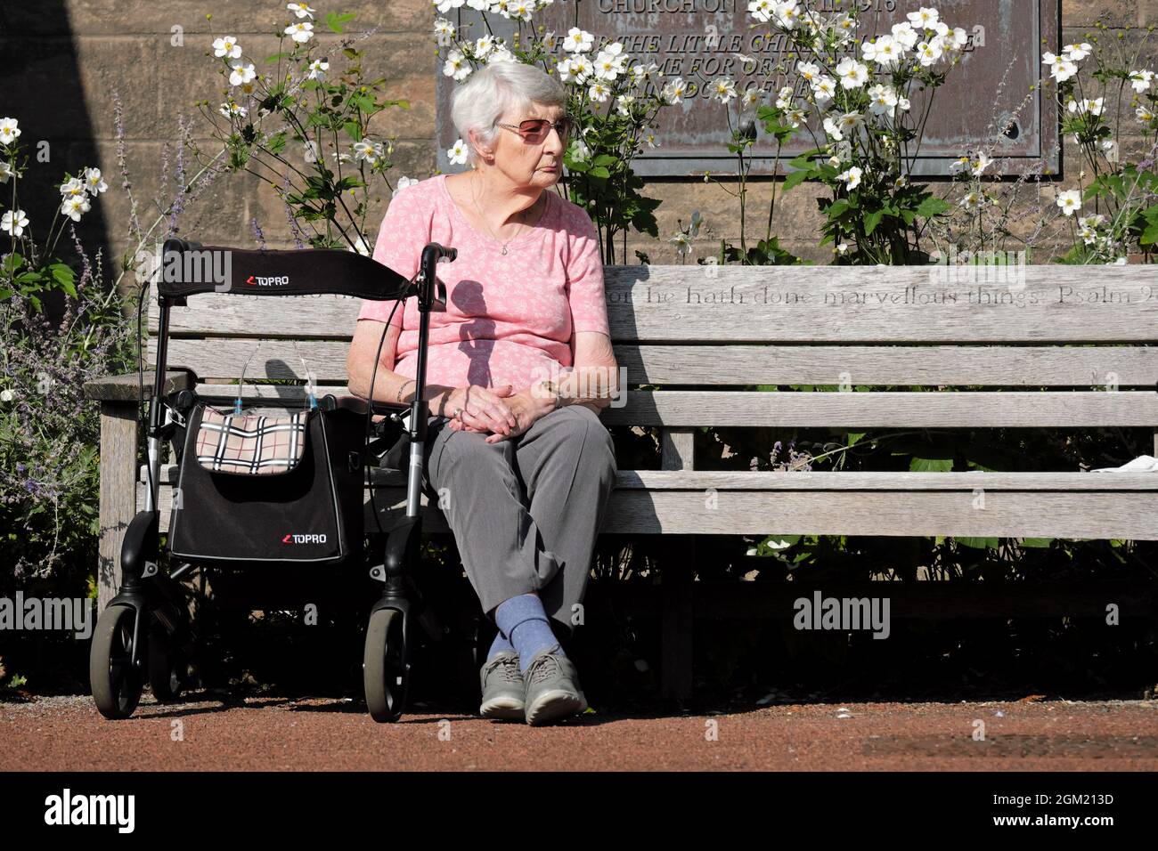 Ältere Frau saß auf der Kirchenbank in der Sonne mit einem Gehrahmen Hilfe 2021. September Großbritannien Stockfoto
