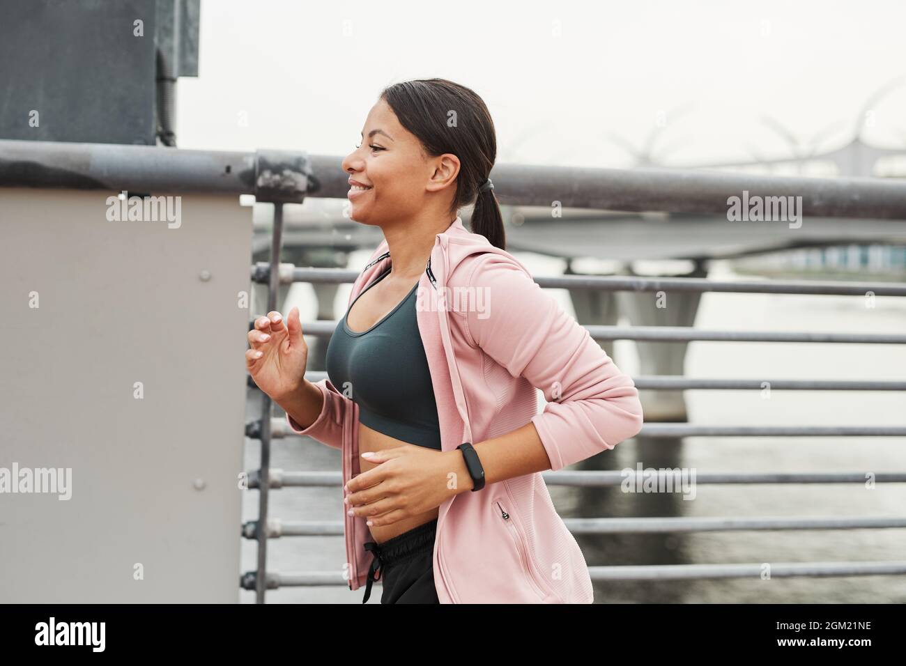 Junger lächelnder Athlet in Sportkleidung, der morgens im Freien entlang der Brücke läuft Stockfoto