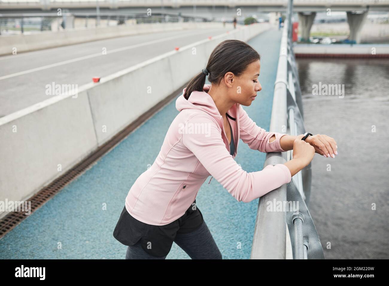 Junge Sportlerin, die auf der Brücke steht und ihren Puls am Fitnessarmband beim Laufen im Freien untersucht Stockfoto