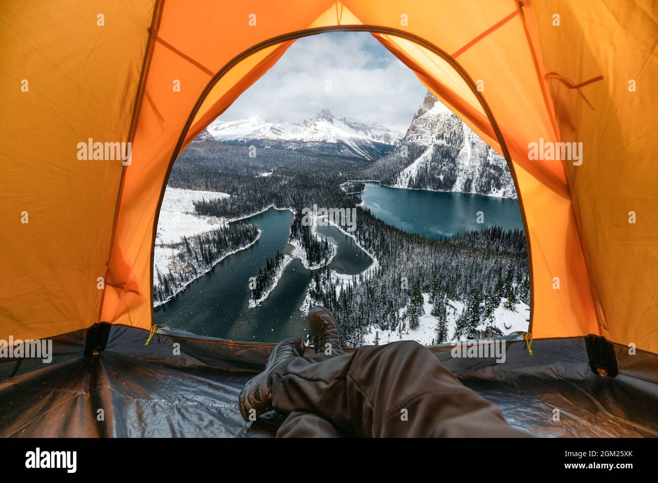 Reisender Mann ruht sich aus und genießt den Blick auf verschneite Berge und See in einem orangefarbenen Zelt im Winter im Yoho Nationalpark, Kanada Stockfoto