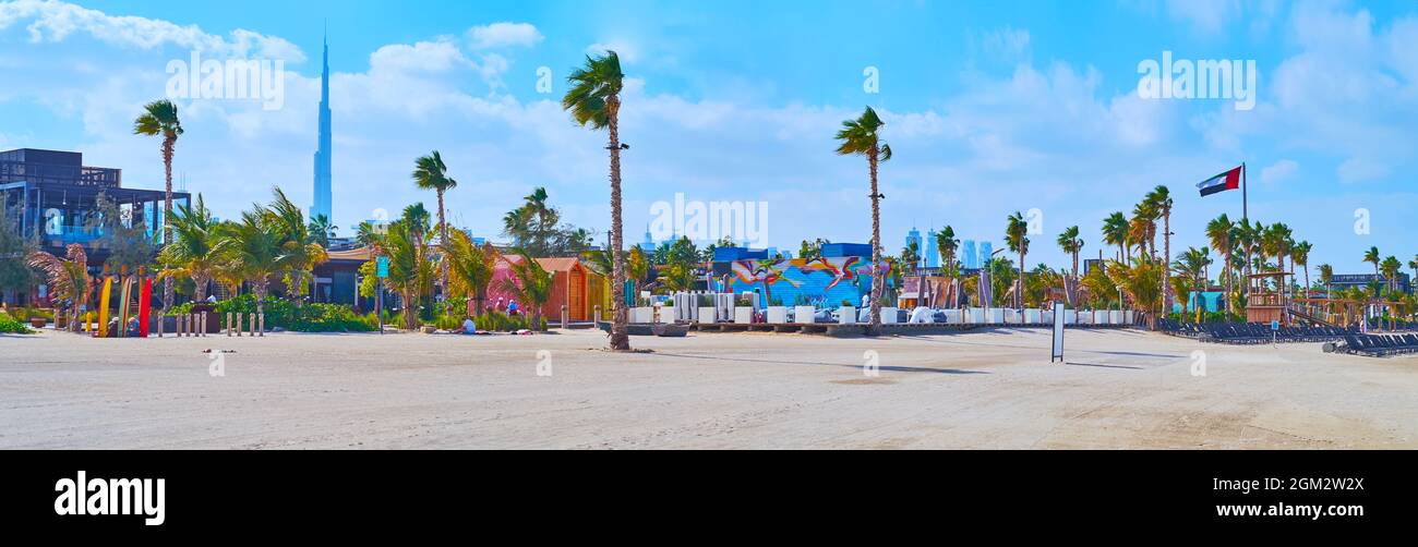 Panorama von La Mer weißen Sandstrand mit einer Reihe von Palmen, Strandläden, Cafés und die Silhouette des Burj Khalifa im Hintergrund, Dubai, VAE Stockfoto