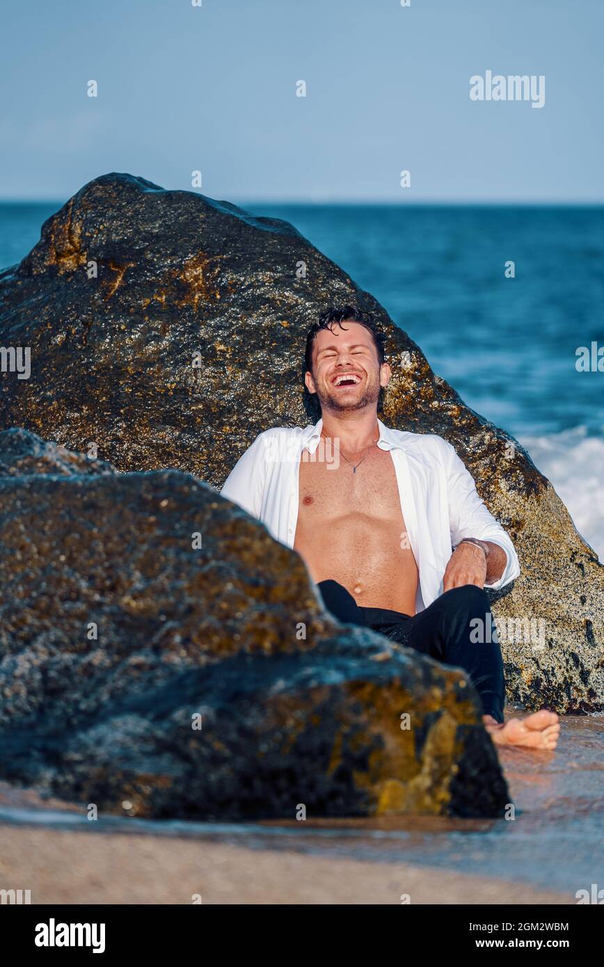 Entzückende trendige Männer in nasser Kleidung, die am Strand in der Nähe von Felsen sitzen und lachen, während sie den Sommer genießen Stockfoto