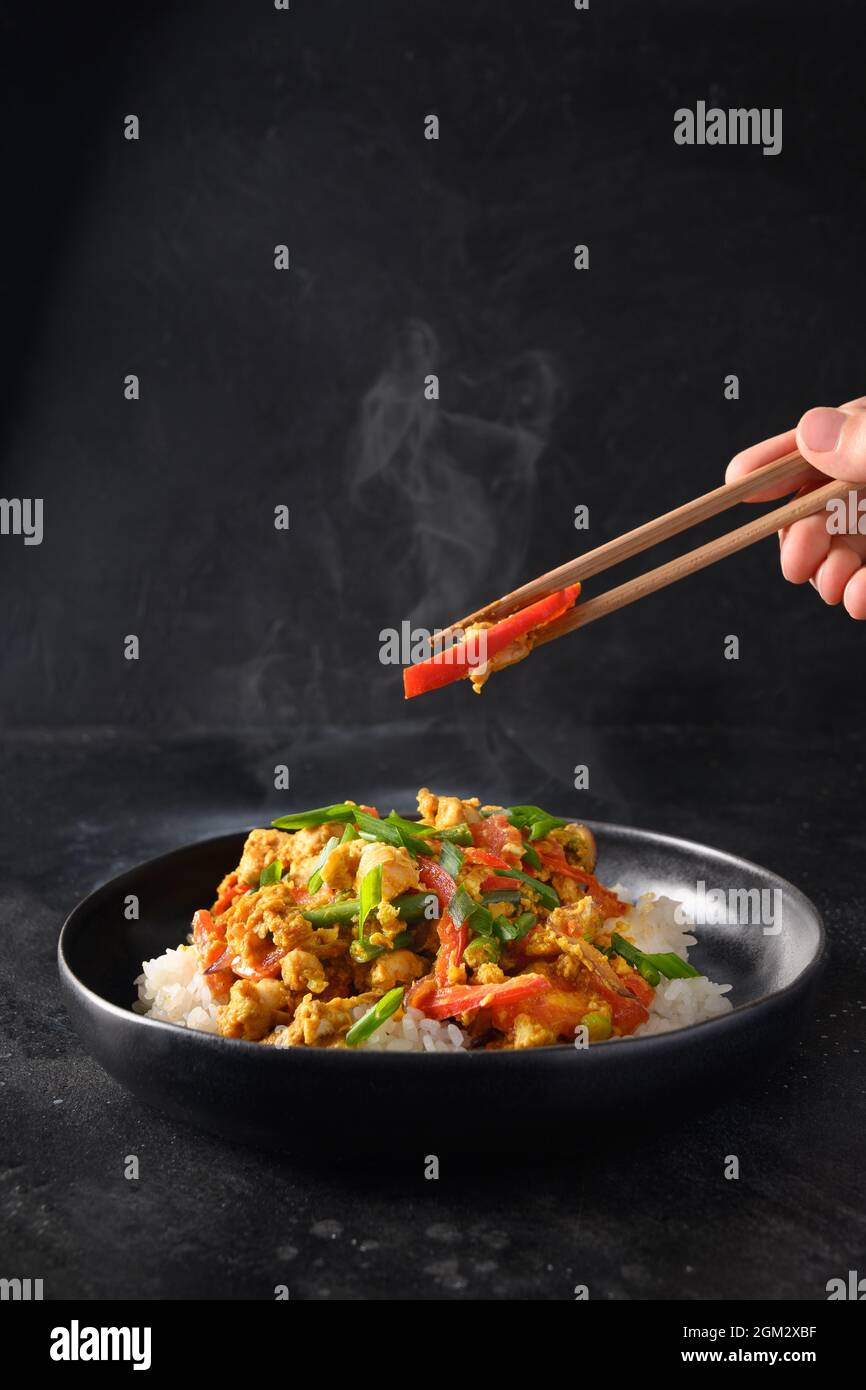 Thailändisches Gericht Gai Pad Pongali mit Huhn, Eiern, würziger, gelber thai-Paprikapaste, Tomaten, Reis in schwarzer Schüssel im Wok. Nahaufnahme. Traditionelle lokale Pause Stockfoto