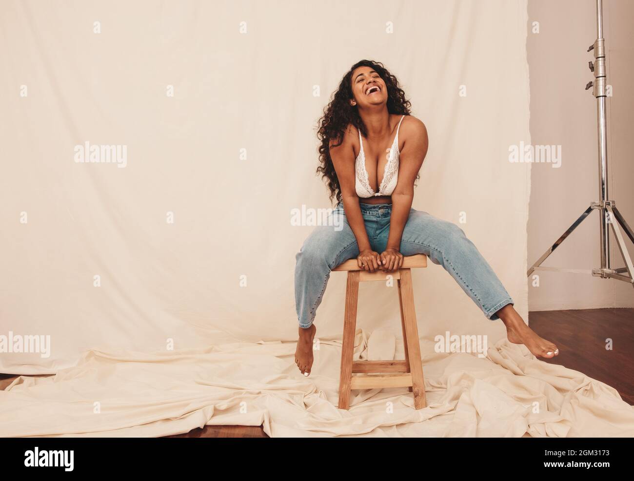 Frau lacht fröhlich, während sie auf einem Stuhl in einem Studio sitzt. Fröhliche junge Frau in Denim-Jeans und einem BH vor einem Studiohintergrund. Junge Wom Stockfoto