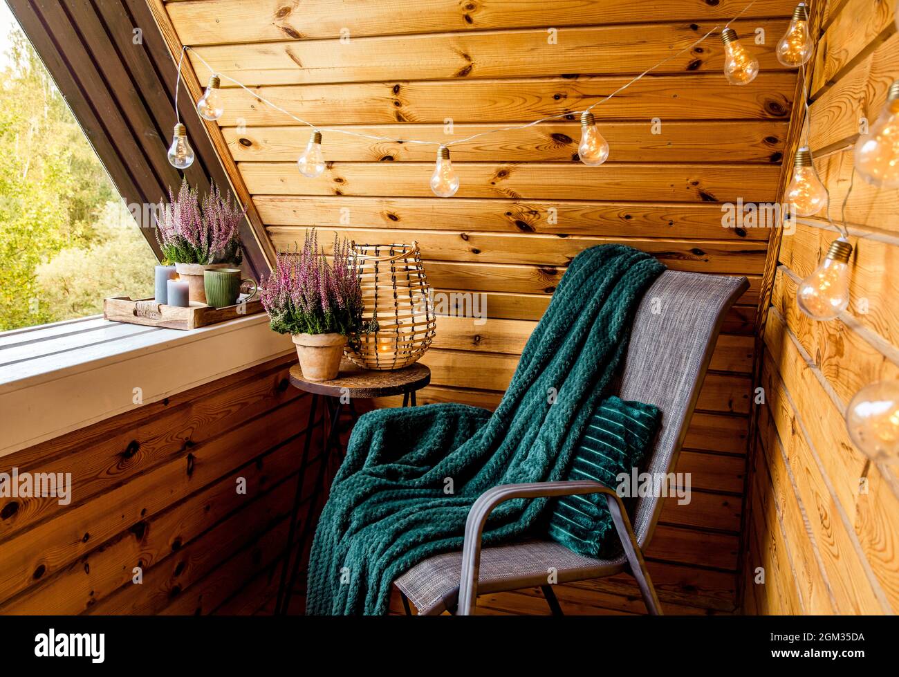 Kleiner, naturfarbener Holzbalkon mit Heideblumen, Kerzenlicht-Flamme, sanftem dunkelgrünem Karli, der auf einem Gartenmöbelstuhl wartet. Stockfoto