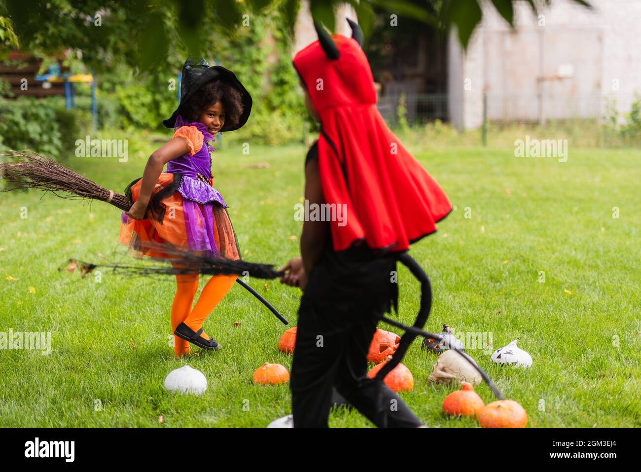 afroamerikanische Geschwister in halloween-Kostümen halten Besen und spielen auf dem Rasen Stockfoto