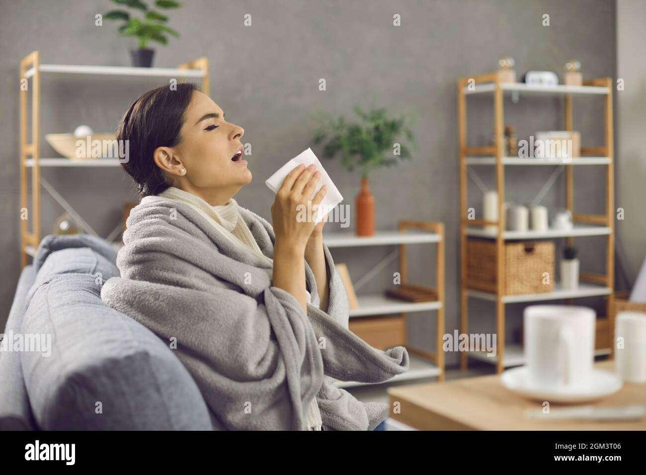 Frau mit einer Serviette in den Händen niest wegen einer laufenden Nase oder Allergie, die zu Hause sitzt. Stockfoto