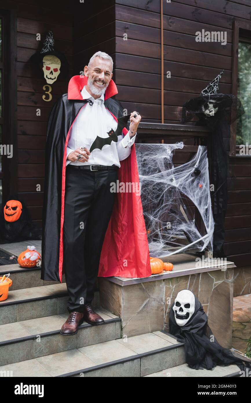 Glücklicher reifer Mann in Vampir Kostüm hält Papier geschnitten Fledermaus  auf Veranda mit halloween-Dekoration Stockfotografie - Alamy