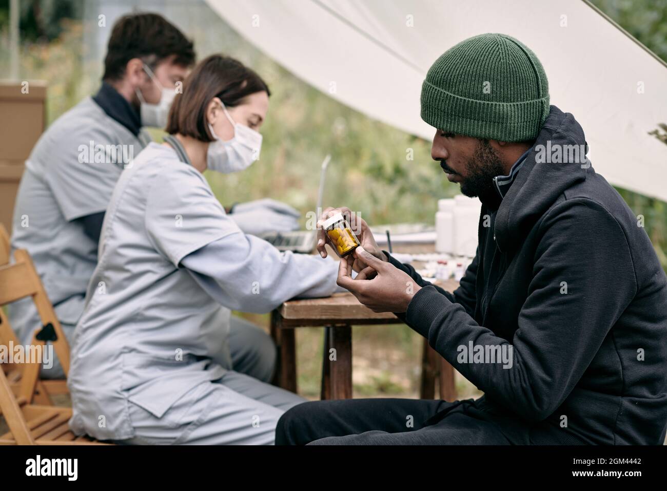 Serious junge schwarze Flüchtling Mann im Hut sitzt am Tisch der Mitarbeiter im Gesundheitswesen und lesen Tag der Pille Flasche, während sie kostenlose Medizin Stockfoto
