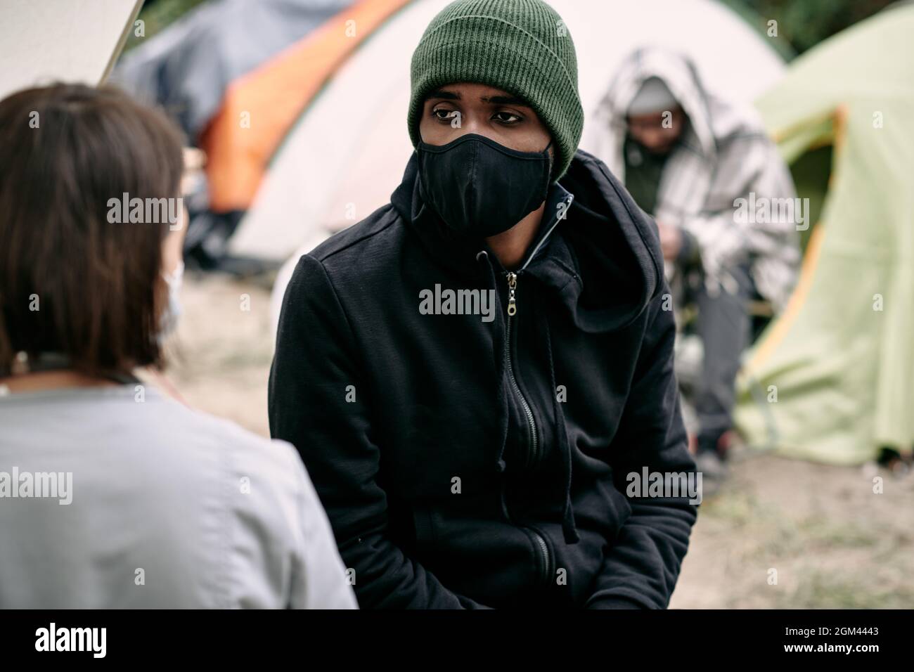 Kranker junger schwarzer Flüchtling in Gesichtsmaske und Hut im Gespräch mit Krankenschwester im Migrantenlager Stockfoto