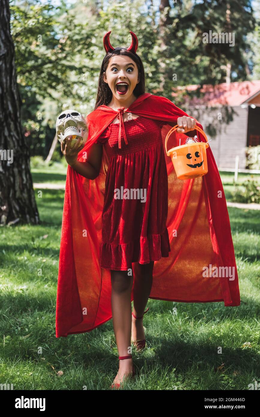 Staunendes Mädchen im dämonischen halloween Kostüm, das mit Totenkopf und  Eimer Süßigkeiten im Freien steht und schreit Stockfotografie - Alamy