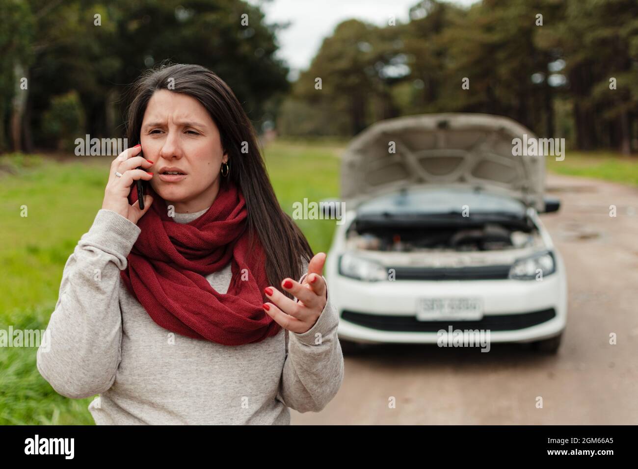 Nahaufnahme einer Latina-Frau, die am Handy spricht und gestikuliert. Ihr kaputtes Auto im Hintergrund Stockfoto