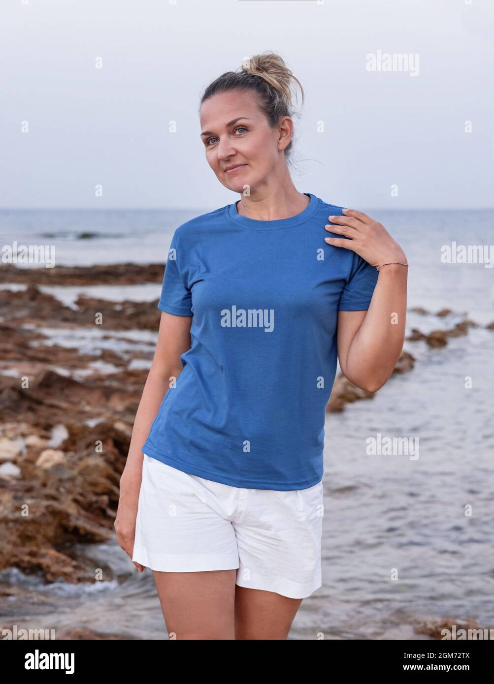 Frau mittleren Alters trägt ein blaues T-Shirt, die Hand auf der Schulter hält und bei Sonnenuntergang auf der Klippe steht. T-Shirt-Modell Stockfoto
