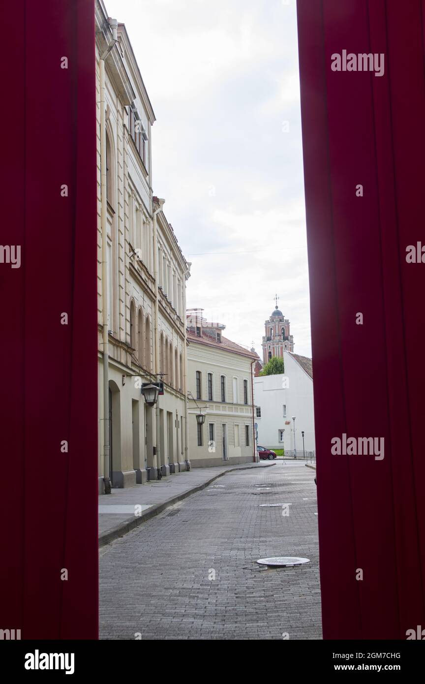 Konzeptionelles Foto der Straße mit Kirche hinter rotem Vorhang in der Altstadt. Architektur von weißen Vintage-Häusern, Apartments mit Fenstern, Balkonen, Boutiquen gegen den bewölkten Himmel im Zentrum von Vilnius Stockfoto