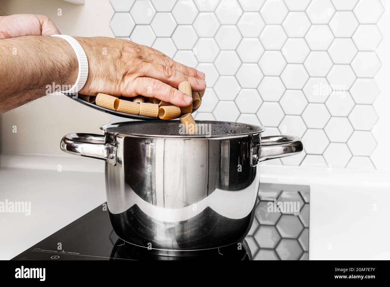 Hände eines erwachsenen Mannes, der italienische Pasta in einen Topf mit kochendem Wasser auf einem Induktionskochfeld gießt Stockfoto