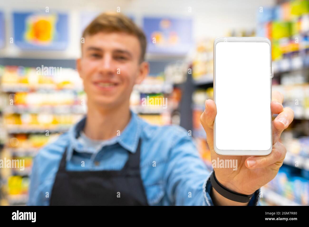 Verschwommenes Foto eines lächelnden Mannes im Geschäft, der eine schwarze Schürze und eine Digitaluhr auf der Hand trägt und die weiße Farbe des Smartphones zeigt Stockfoto