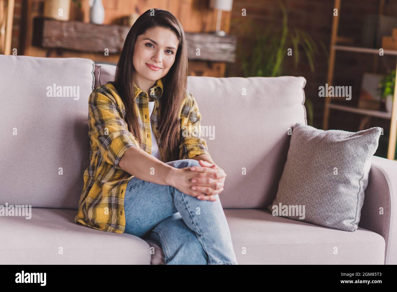 Foto von ruhigen verträumt niedlich charmante Dame aussehen Kamera Umarmung Knie sitzen Sofa tragen karierte Hemd zu Hause drinnen Stockfoto
