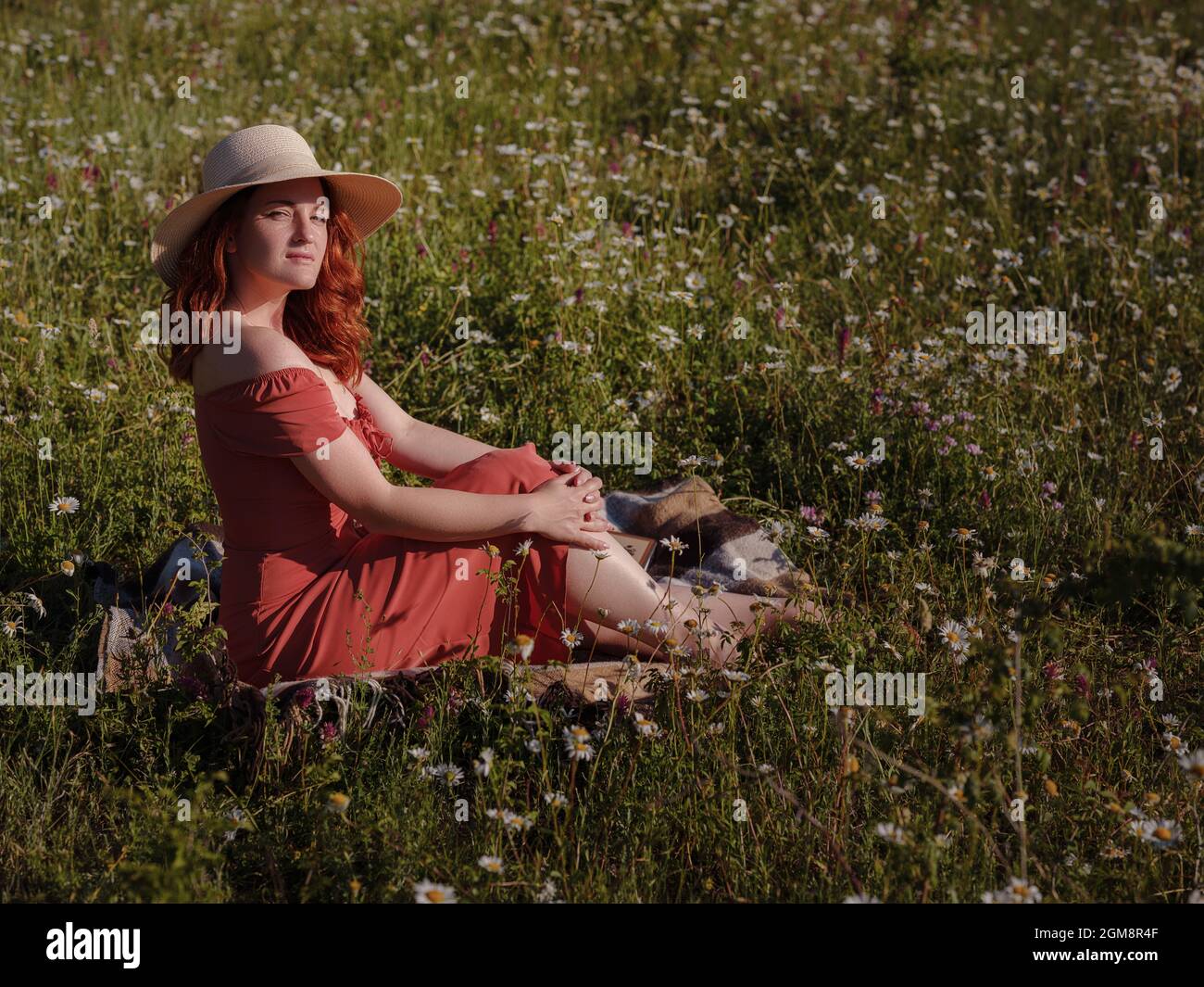 Rothaarige Frau trägt Kleid und Hut auf Sommerabend Feld der Gänseblümchen Lesen eines Buches, während sie auf einer Decke sitzen . Konzept der digitalen Entgiftung, Digita Stockfoto